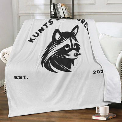 White soft cotton blanket with black rebellious raccoon graphic and text "KUNTS" and "EST. 2023", styled in punk streetwear design