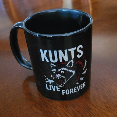 Black coffee mug with aggressive raccoon graphic and text "KUNTS LIVE FOREVER" on a wooden surface